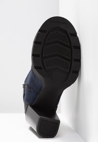 Botte à talon avec semelle noire, semelle extérieure texturée et dessus en tissu marine, positionnée semelle vers le haut contre un mur blanc d’angle.