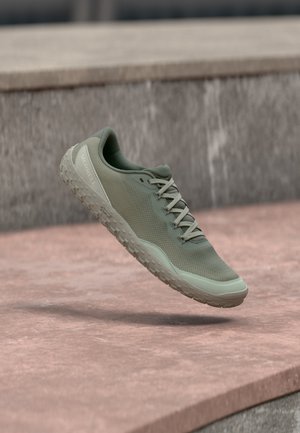 Chaussure de sport Merrell vert clair avec semelle texturée et lacets, suspendue au-dessus d'une surface en pierre rose et d'un fond en pierre grise.