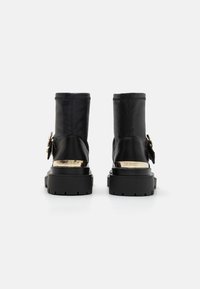 Botas de cuero negras hasta el tobillo con una suela gruesa negra, detalles metálicos dorados en el tacón y un detalle decorativo de hardware en el costado.