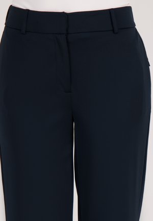 Close-up van zwarte op maat gemaakte broeken met riemlussen en een ritssluiting aan de voorkant, gedragen door iemand in een lichtroze top.