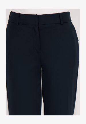 Close-up van zwarte op maat gemaakte broeken met riemlussen en een ritssluiting aan de voorkant, gedragen door iemand in een lichtroze top.