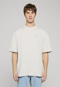 Světlé béžové oversized bavlněné tri�čko s kulatým výstřihem a krátkými rukávy. Na hrudi má jemné logo. Jednoduchý design.