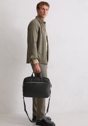 Homme en chemise et pantalon beige tenant une mallette noire avec une bandoulière, debout contre un mur blanc.