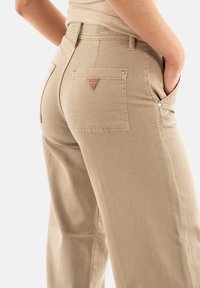 Pantalones de algodón de pierna ancha en color beige con un bolsillo trasero que presenta un logo de triángulo rojo, remaches metálicos en los bolsillos y un acabado de tejido texturizado.