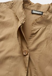 Camicia tan con colletto arricciato, chiusura con un bottone e texture liscia, con un'etichetta che mostra "Blauer" all'interno.