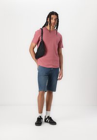 Rode korte mouwen t-shirt, spijkerbroek shorts, zwarte crossbody tas met band, witte sokken, zwarte sneakers met witte accenten, staand tegen een witte achtergrond.