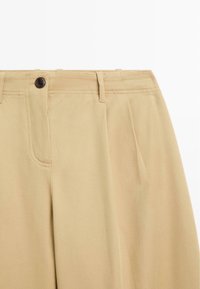 Beige Hochbundhosen mit Gürtelschlaufen, Knopfverschluss und Falten auf der Vorderseite, aus einem strukturierten Stoff gefertigt.