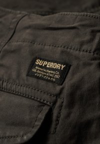 Écusson en tissu noir arborant le logo "SUPERDRY" en doré, avec du texte en dessous. La texture est lisse ; les bords sont soigneusement cousus avec une poche visible.