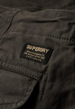 Zwarte stoffen patch met het "SUPERDRY" logo in goud, met tekst eronder. De textuur is glad; de randen zijn netjes gestikt met een zichtbare zak.
