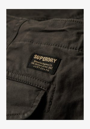 Toppa di tessuto nero con logo "SUPERDRY" in oro, con testo sottostante. La trama è liscia; i bordi sono cuciti con cura e c'è una tasca visibile.