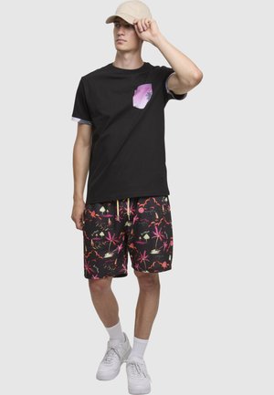 Zwart T-shirt met een paars zakje met palmbladpatroon, gecombineerd met kleurrijke zwarte shorts met roze en oranje tropische motieven. Witte sneakers.
