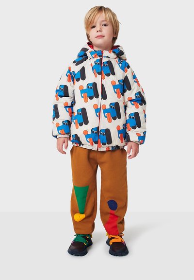 Chaqueta blanca esponjosa con gráficos coloridos de personajes abstractos, combinada con pantalones marrones que presentan parches verdes, rojos y azules. Zapatos negros.