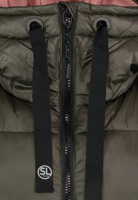 Gros plan sur l’avant d’une veste matelassée vert foncé montrant une fermeture éclair noire et deux cordons noirs avec le logo SL Studio à l’extrémité gauche du cordon.
