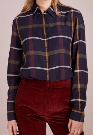Persoon draagt een donkerblauw geruit overhemd met knopen met oranje, kastanjebruine en groene strepen, ingestopt in een hoge taille dieprode ribfluwelen broek.