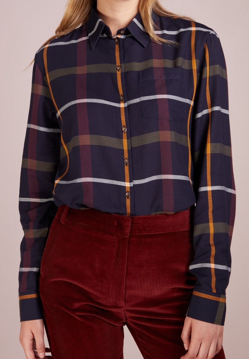 Personne portant une chemise à carreaux bleu marine foncé à boutons avec des rayures orange, bordeaux et vertes, rentrée dans un pantalon en velours côtelé rouge profond à taille haute.
