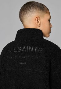 Juoda sherpa striukė su aukštu apykakle, kurios nugaroje išspausta "ALLSAINTS" tekstas ir subtilūs akcentai. Minkšta tekstūra ir laisvas pritaikymas.