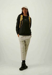 Fiatal nő bézs nadrágban, fekete pulóverben, világosbarna hátizsákkal, fekete sportcipőben, fekete zokniban és bézs sapkában áll semleges háttér előtt.