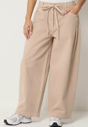 Kvinde iført beige bukser med vide ben og en bindebælte i taljen, kombineret med hvide og beige sneakers, står med den ene hånd i lommen.