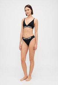 Crni bikini set s braletom na V-izrezu i visokim donjim dijelom, s bijelim Moschino logotip trakom. Glatka, rastezljiva tkanina.