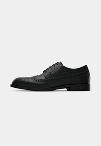Chaussure habillée pour homme en cuir noir avec détails brogue et lacets, vue de côté sur fond clair uni.
