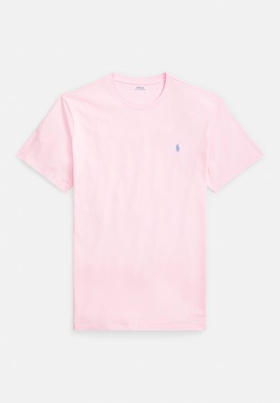 Rožinė medvilninė marškinėliai su apvalia iškirpte, trumpomis rankovėmis ir mažu mėlynu logotipu ant krūtinės. Lygus tekstūros ir klasikinio stiliaus.