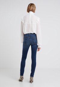 Blusa blanca con mangas acampanadas, espalda texturizada; combinada con jeans oscuros ajustados y zapatos de tacón con estampado. Fondo neutro.