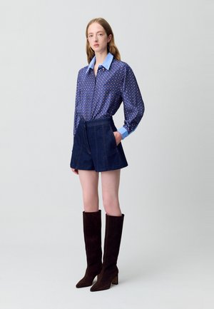 Camisa de manga larga azul con lunares, combinada con pantalones cortos de mezclilla oscuros y botas de ante marrón hasta la rodilla con tacón cuadrado.