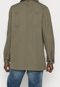 Giacca di tessuto verde oliva con un yoke posteriore, quattro bottoni a toggle sui polsi e due pattine per le tasche frontali. Texture liscia e resistente.