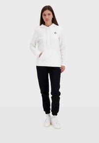 Vit hoodie med en framficka, justerbara dragsnören och en färgglad logotyp på bröstet; kombinerad med svarta joggers och vita sneakers.