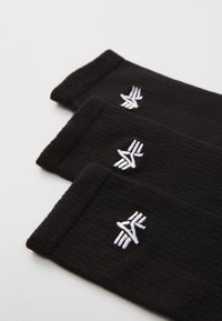 Schwarze Socken mit geripptem Muster, mit einem weißen gestickten Logo im Bereich des Bündchens. Drei Paar horizontal übereinander gestapelt.