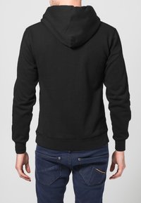 Felpa nera con cappuccio, dalla texture liscia, polsini e fascia in vita a costine, vista da dietro. Indossata sopra jeans di denim scuro con tasche.