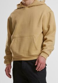 Muž v béžové mikině s kapsou na přední straně a černých kalhotách, stojící před jednolitým světlým pozadím, ruce u těla.