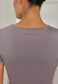 Aktivwear top i dæmpet taupe med en glat tekstur, med rund halsudskæring og ordet "ACTIVE" trykt på ryggen.