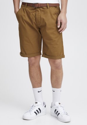 Khakishorts mit umgeschlagenen Säumen, seitlichen Taschen und einer Vorderfalte, kombiniert mit weißen Sneakern und Sportsocken mit einem schwarzen Logo.