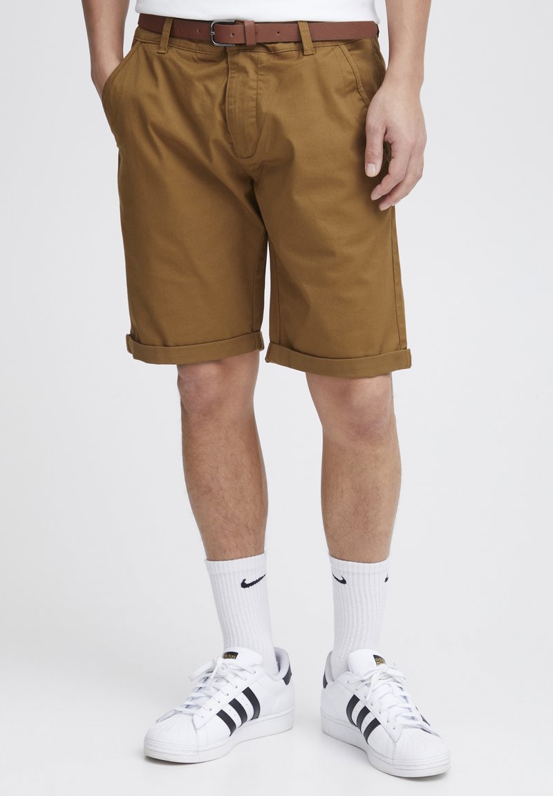 Khaki shorts with rolled cuffs, side pockets, and a front pleat, paired with white sneakers and athletic socks featuring a black logo.