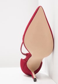 Röd mocka högklackad sko med spetsig tå och ankelrem, sålen vänd framåt mot vit hörnbakgrund.