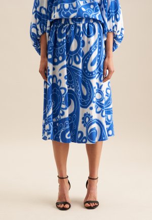 Jupe bleue et blanche avec un motif paisley, taille élastique, mi-longue, accompagnée de sandales noires à lanières. Texture de tissu lisse.