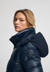 Doudoune marine à texture brillante, col montant et capuche en tissu contrasté. Présente un patch logo sur la manche gauche.