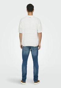 Hvit kortermet strikket skjorte med teksturert mønster, kombinert med lys blå denim jeans og brune joggesko, sett fra baksiden.