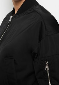 Chaqueta bomber negra con cierre de cremallera, cuello acanalado y dos bolsillos laterales. Presenta una tela suave y detalles de costuras visibles.