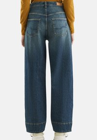 Jeans o wysokim stanie i szerokich nogawkach w ciemnoniebieskim dżinsie, z eleganckim wykończeniem, tylnymi kieszeniami i mankietami na końcu nogawek, w połączeniu z białymi sneakersami.