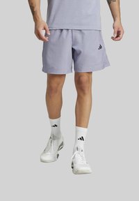 Man in een lichtpaarse Adidas-short, witte Adidas-sokken en witte sportschoenen, staand tegen een effen grijze achtergrond.
