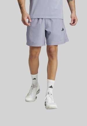 Hombre vestido con pantalones cortos Adidas de color púrpura claro, calcetas Adidas blancas y zapatillas deportivas blancas, de pie contra un fondo gris liso.