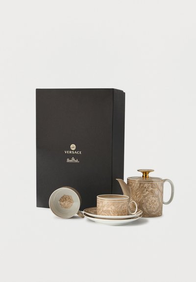 Luxury Versace tea set with gold and beige ornate patterns, including teapot, cup, saucer, and lid, beside a black Versace gift box.