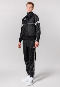 Sort træningsdragt lavet af glat stof, med tre hvide striber på jakken og bukserne, og parret med hvide sneakers.