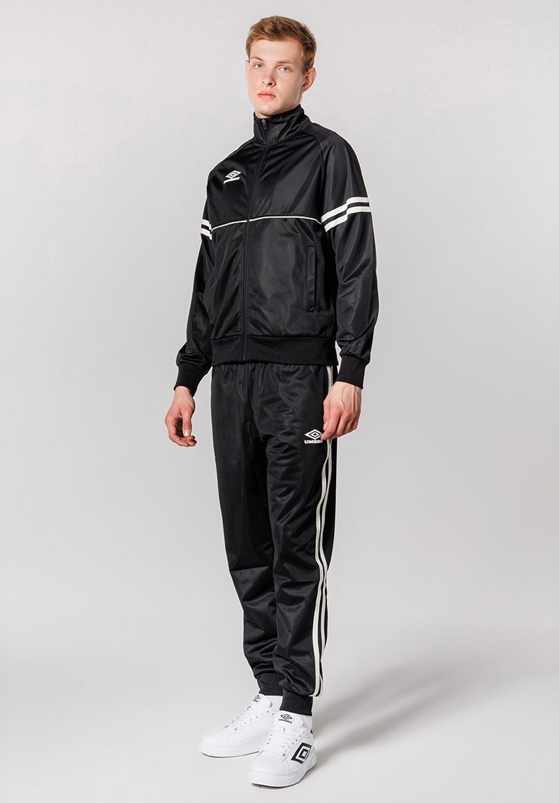 Sort træningsdragt lavet af glat stof, med tre hvide striber på jakken og bukserne, og parret med hvide sneakers.