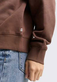 Brązowy sweatshirt z miękkiej bawełny z ściągaczami u dołu, z przeszytym wykończeniem i małą metką logo. Zestawiony z jasnoniebieskimi dżinsami.