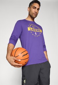 Lilla langærmet trøje med "LAKERS" logo, sorte shorts, orange basketball i hånden. Tekstureret stof, atletisk pasform, sporty design.