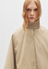 Cappa impermeabile beige con colletto alto, caratterizzata da un design minimalista e una texture liscia. Il capo ha una forma ampia e fluida.