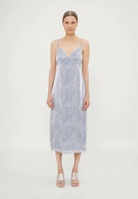 Žena s punđom, obučena u bezrukavnu, svijetloplavu midi haljinu s uzorkom i srebrnim sandalama s otvorenim prstima, stoji protiv jednostavne pozadine.